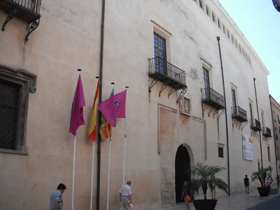 Fachada de piedra del Palacio Ducal de los Borja en Gandia, con balcones de hierro, banderas y una portada de arco de medio punto