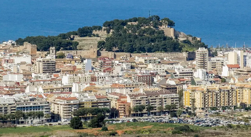 Castillo de Dénia sobre una colina con pinos en medio de la ciudad, con el puerto y el mar detrás, visto desde el Montgó