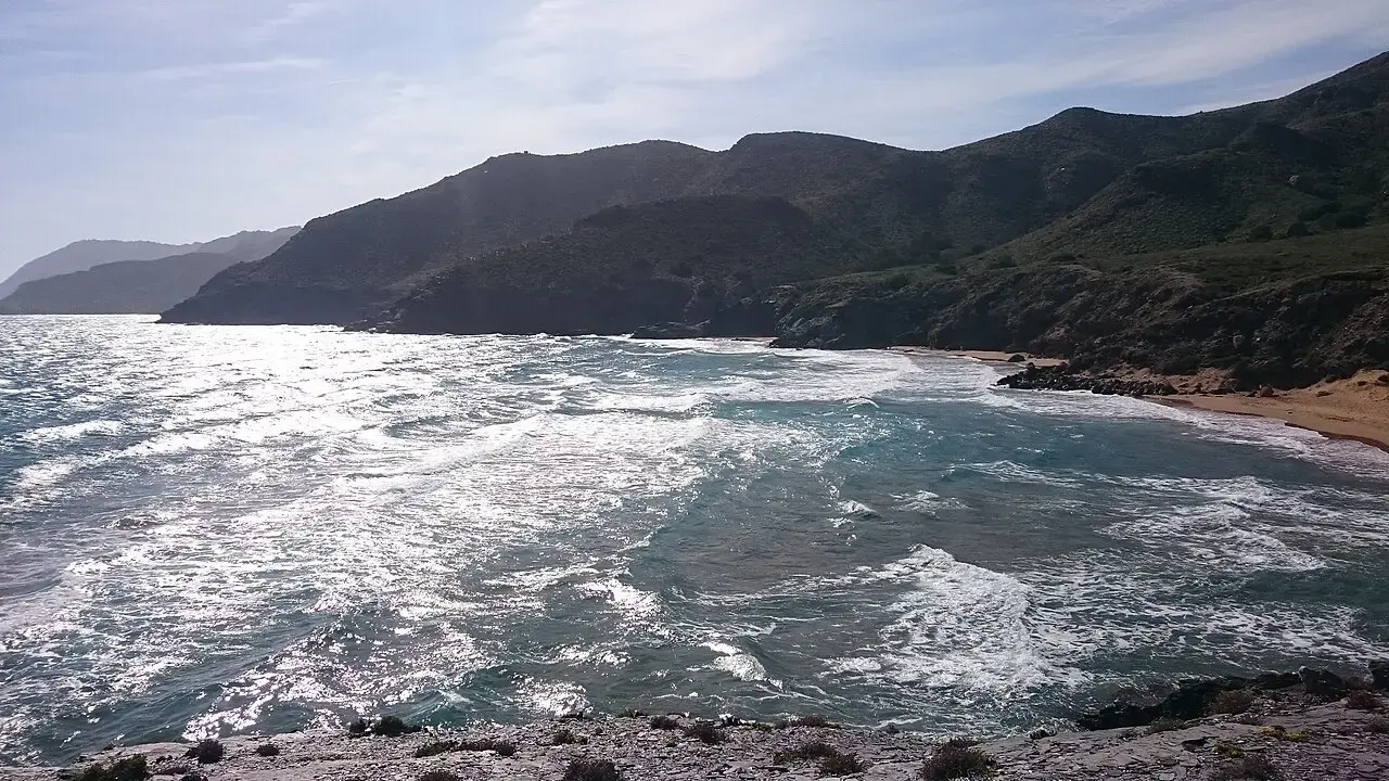 Costa de Calblanque con oleaje y espuma bajo el sol, una playa de arena dorada y montes oscuros al fondo