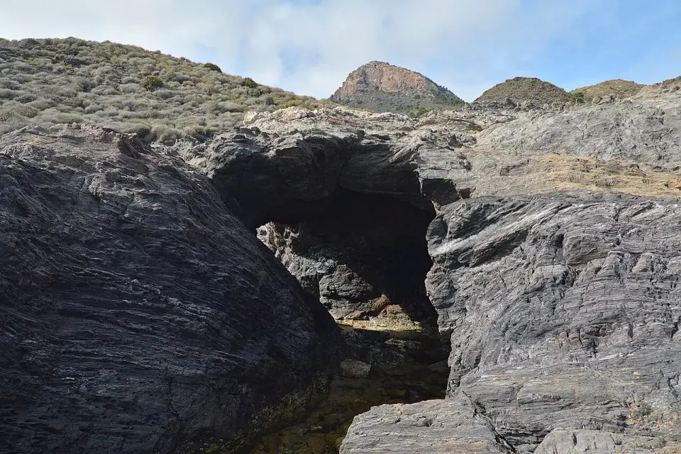 Rocas oscuras con una pequeña cueva en la costa de Calblanque, con un monte puntiagudo al fondo