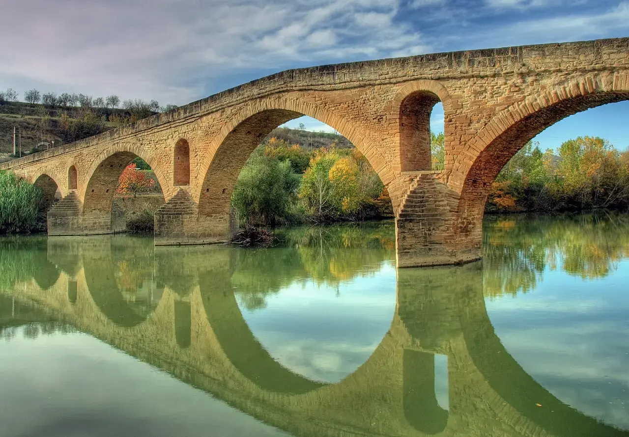 Puente románico de Puente la Reina con sus arcos de piedra reflejados en el río Arga, entre árboles con colores de otoño