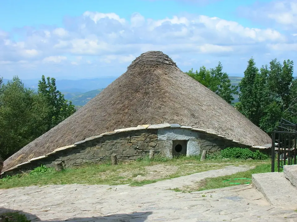 Palloza de piedra con tejado de paja en O Cebreiro, con montañas y bosque al fondo