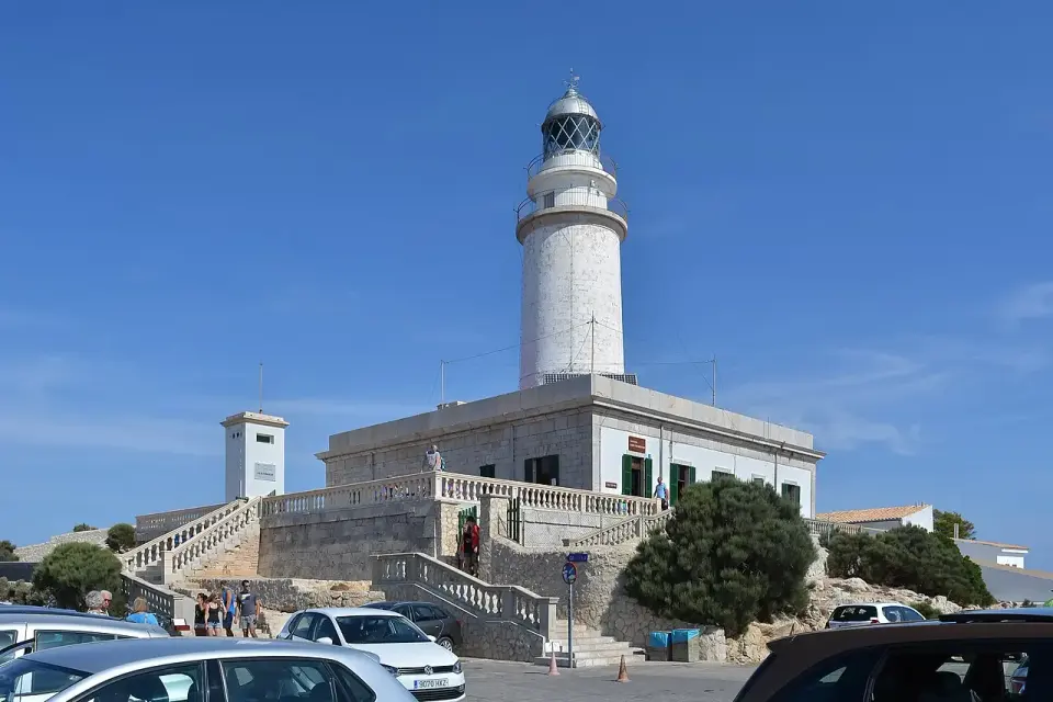 Faro blanco del Cap de Formentor sobre su edificio de piedra, con coches aparcados delante