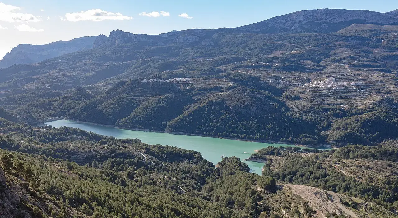 Embalse de Guadalest de aguas turquesas entre montañas y pinares, con el pueblo en lo alto de la ladera