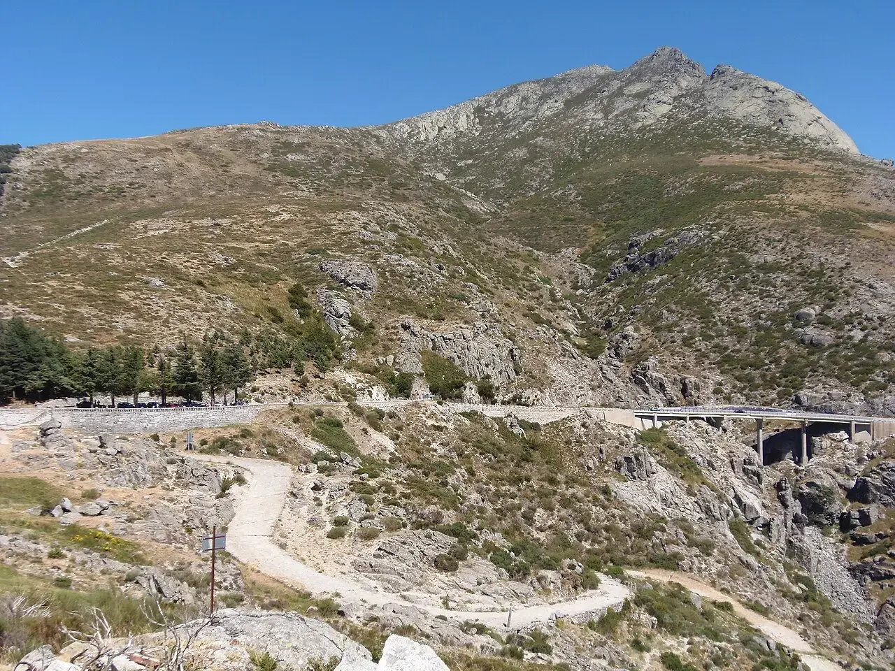 Calzada empedrada en zigzag bajando por la ladera del puerto del Pico, con el viaducto de la N-502 y las cumbres de Gredos al fondo