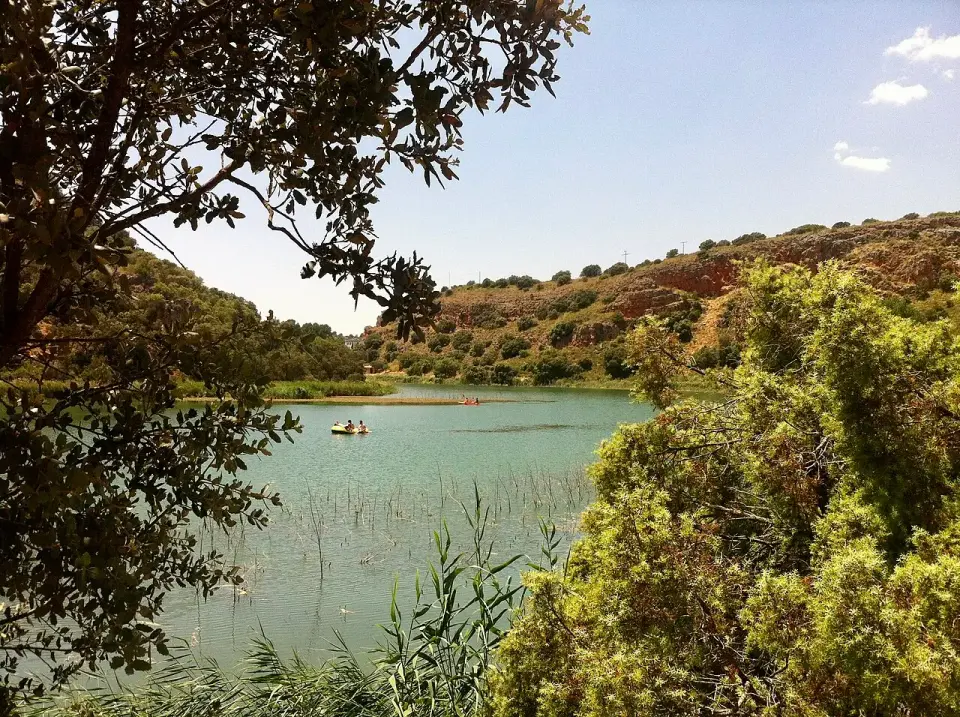 Laguna de San Pedro vista entre las ramas de una encina, con barcas en el agua y laderas de roca rojiza