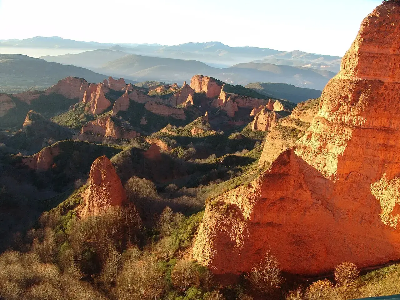 Picachos de arcilla rojiza de Las Médulas iluminados por el sol, entre castaños y con montañas al fondo