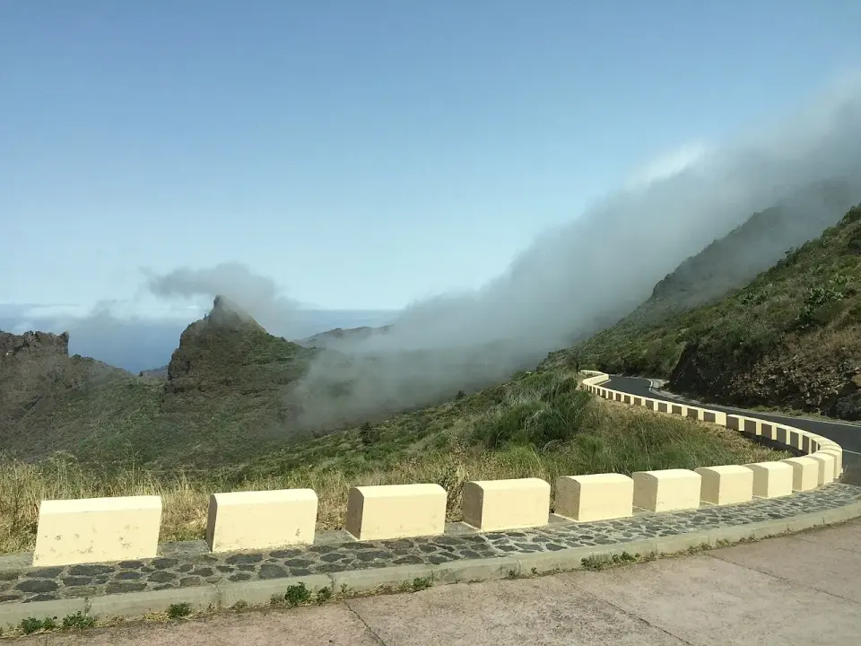 Curva de la TF-436 con bolardos blancos sobre el valle de Masca, con nubes entrando por las montañas y el mar al fondo