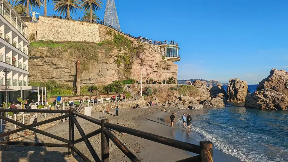 Mirador del Balcón de Europa en Nerja sobre un promontorio rocoso, con palmeras, una playa y paseantes junto al mar