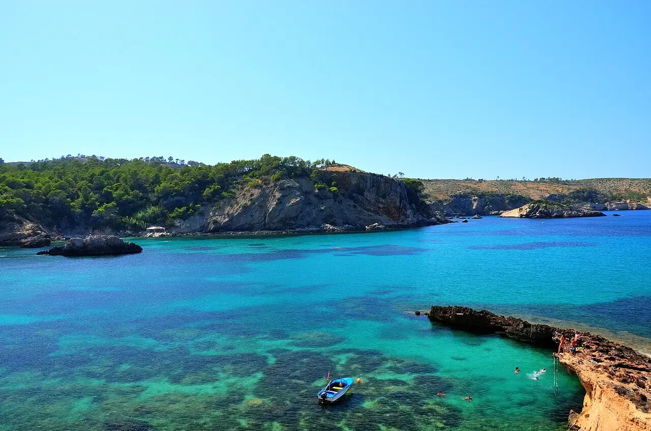 Cala Xarraca, con agua turquesa sobre fondos de roca y una barca, rodeada de acantilados con pinos