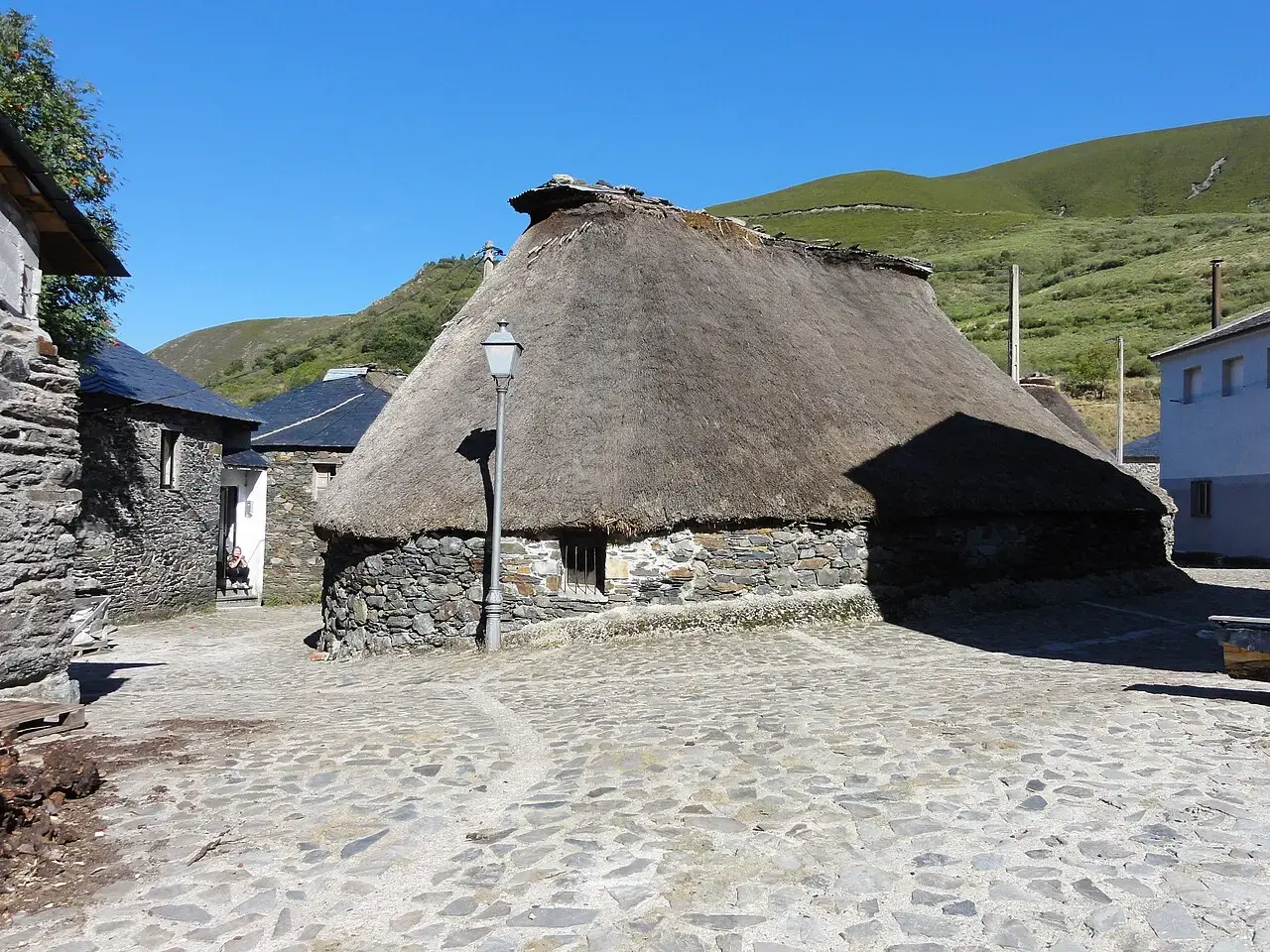 Palloza de piedra con tejado de paja junto a una calle empedrada en Balouta, con prados de montaña al fondo