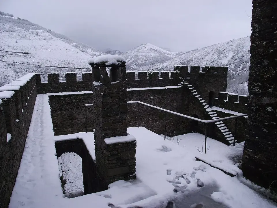 Patio y murallas almenadas del castillo de Doiras cubiertos de nieve, con montañas nevadas detrás