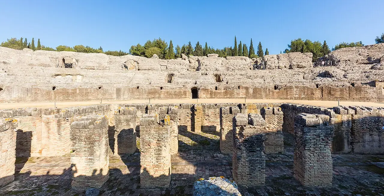 Arena y graderíos de piedra del anfiteatro romano de Itálica, con la fosa central y pinos al fondo bajo un cielo azul