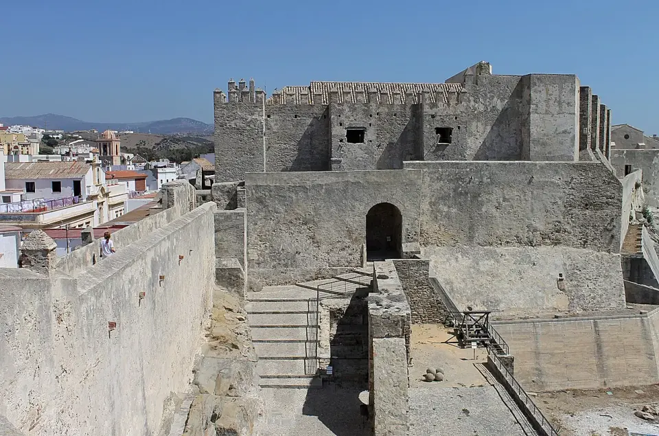 Patio de armas y murallas de piedra del castillo de Guzmán el Bueno en Tarifa, con tejados del pueblo y montes al fondo