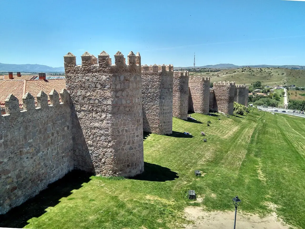 Lienzo de la muralla de Ávila con sus cubos almenados de granito sobre un talud de césped verde y el campo al fondo