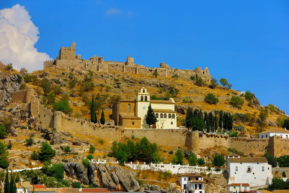 Murallas y castillo de Moclín en lo alto de un cerro de matorral, con una iglesia blanca en la ladera y cielo azul