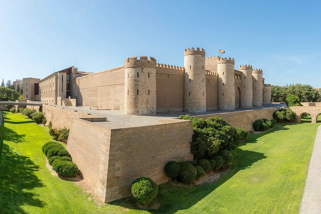 Murallas de piedra con torreones del palacio de la Aljafería de Zaragoza, rodeadas de un foso ajardinado con césped