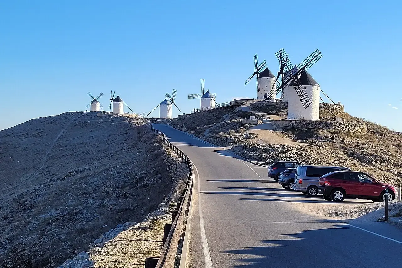 Carretera que sube al cerro Calderico de Consuegra entre molinos de viento blancos, con coches aparcados junto a la cuneta