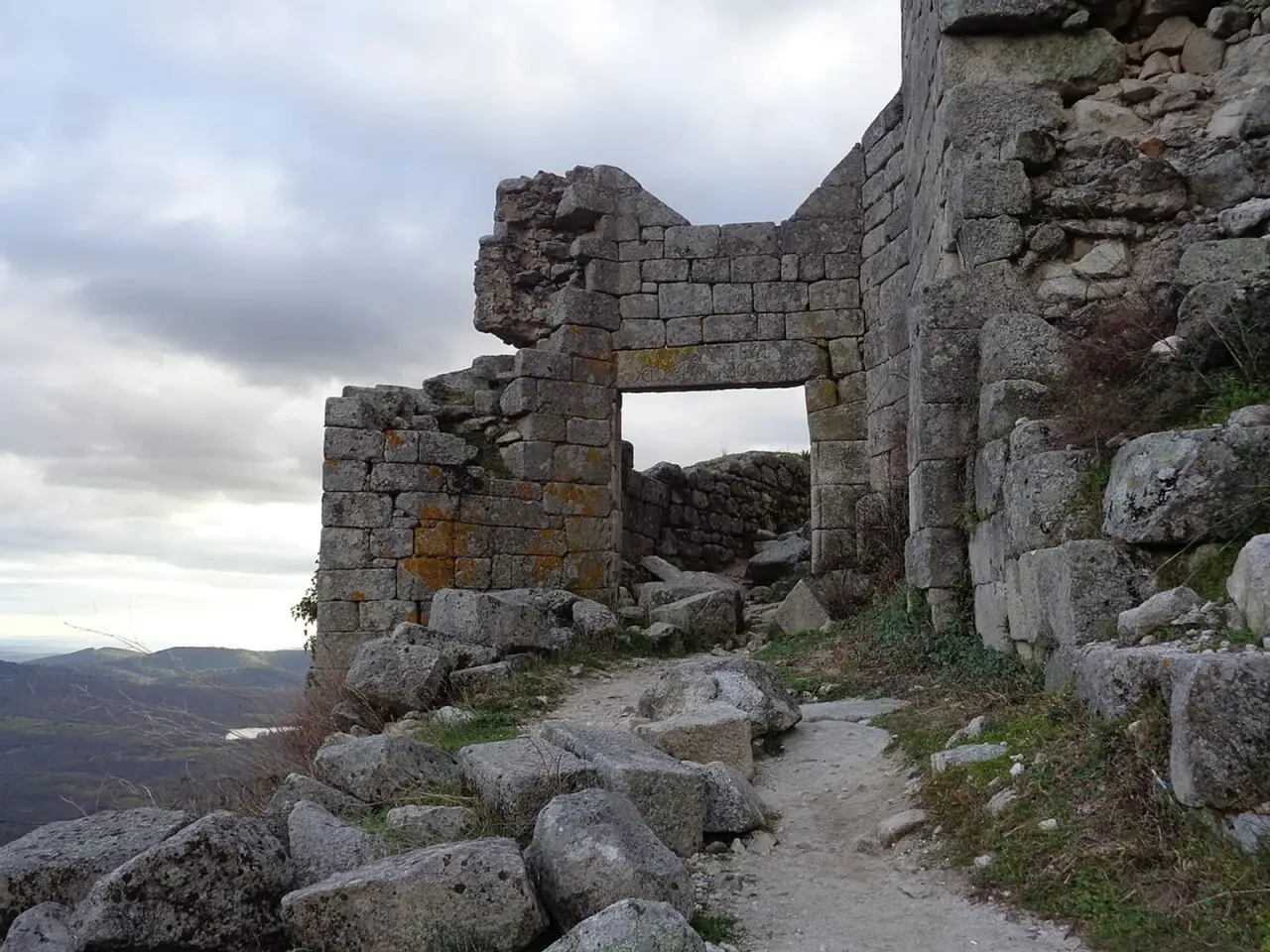 Arco de piedra en ruinas del castillo de Trevejo, con un sendero entre rocas de granito y montañas al fondo