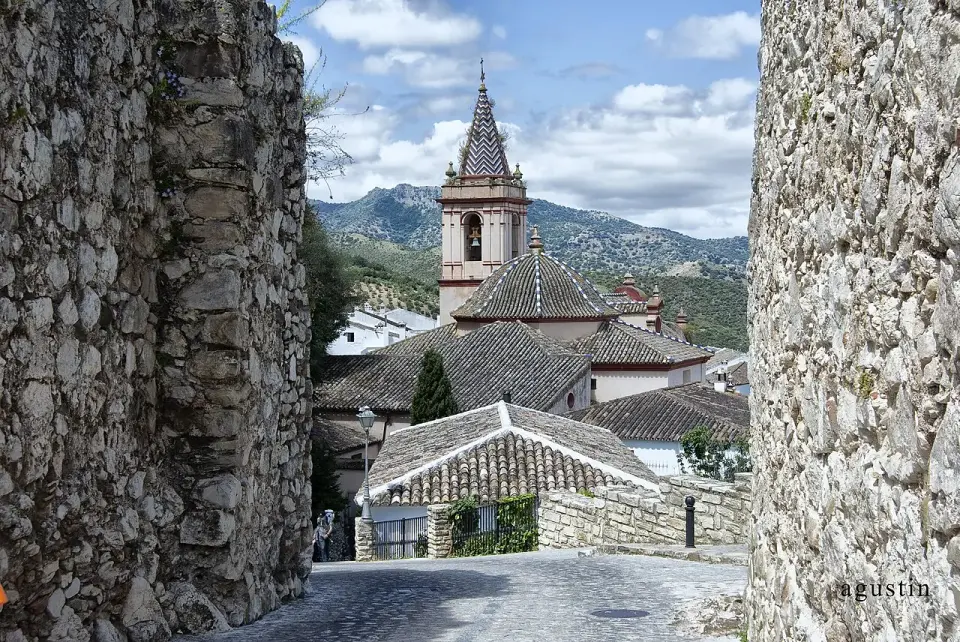 Calle empedrada entre muros de piedra que se abre a la iglesia de Zahara de la Sierra, con la sierra detrás