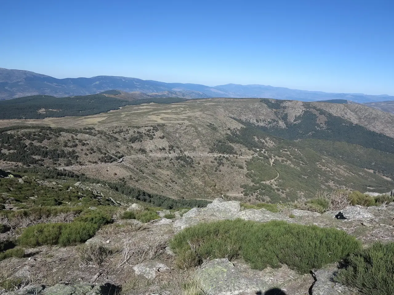 La carretera M-611 subiendo hacia el puerto de la Morcuera entre laderas de matorral y pinar, vista desde lo alto de La Najarra con un cielo azul despejado