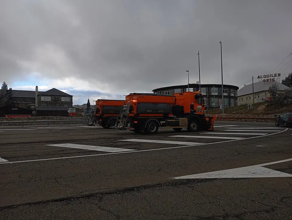 Dos quitanieves naranjas aparcados en la explanada del puerto de Navacerrada, con edificios de montaña y un cartel de alquiler de esquís al fondo bajo un cielo nublado
