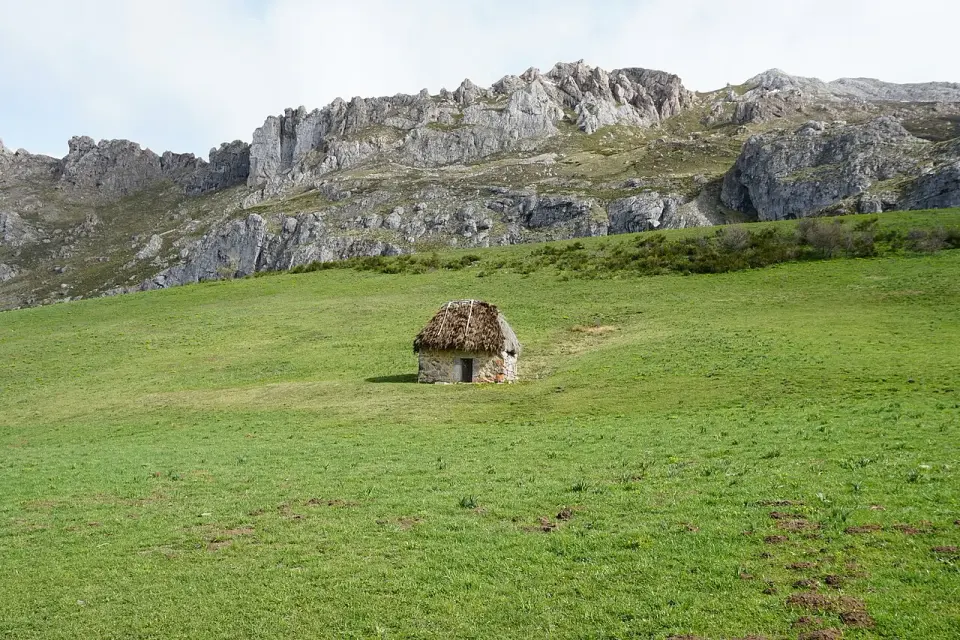 Cabaña de piedra con tejado vegetal, un teito, en medio de una gran pradera de Valle de Lago, con crestas calizas al fondo