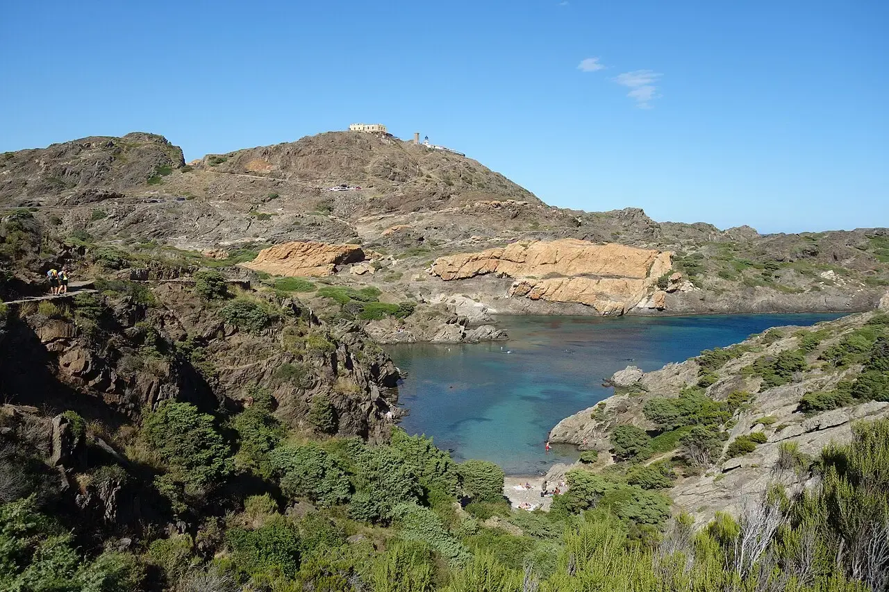 Cala de aguas turquesas entre rocas y matorral en el Cap de Creus, con el faro en lo alto de la colina